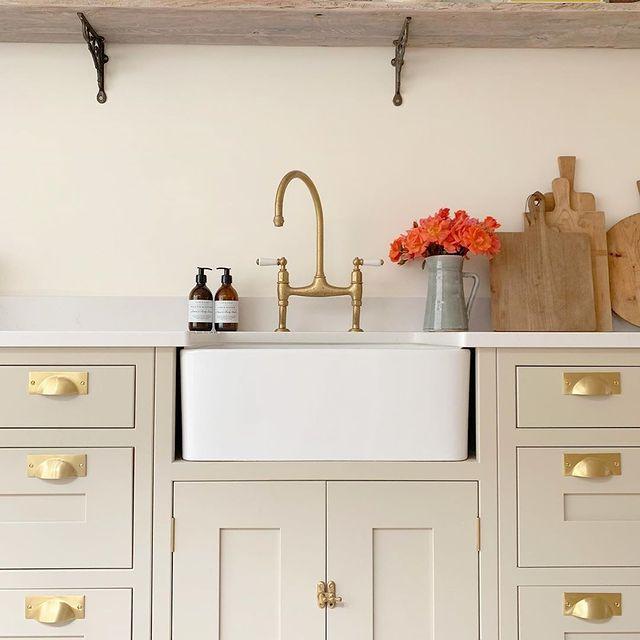 The M.Marcus Heritage Brass Cheshire Cup Handle in Satin Brass on a cream cabinet with a centred white sink | More Handles